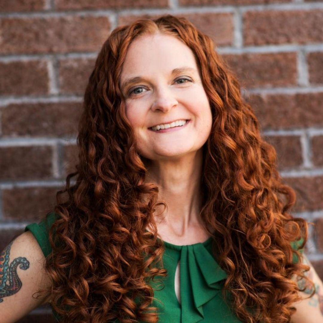 red, curly haired woman in green dress standing in front of a brick wall smiling at the camera
