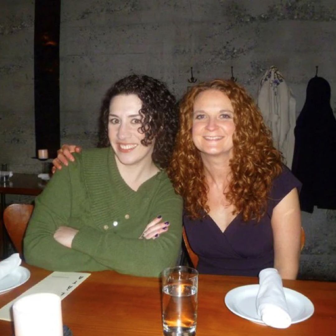 Two curly haired woman sitting at a dinner table, one with their arm around the other