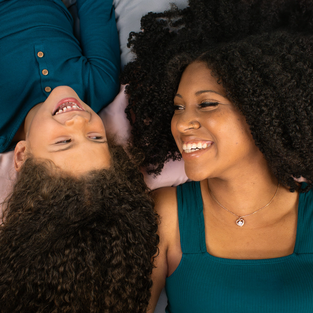 curl haired mother and young daughter laying in bed smiling at each other and each holds a bottle of Awe Inspiraling Refresher Spray