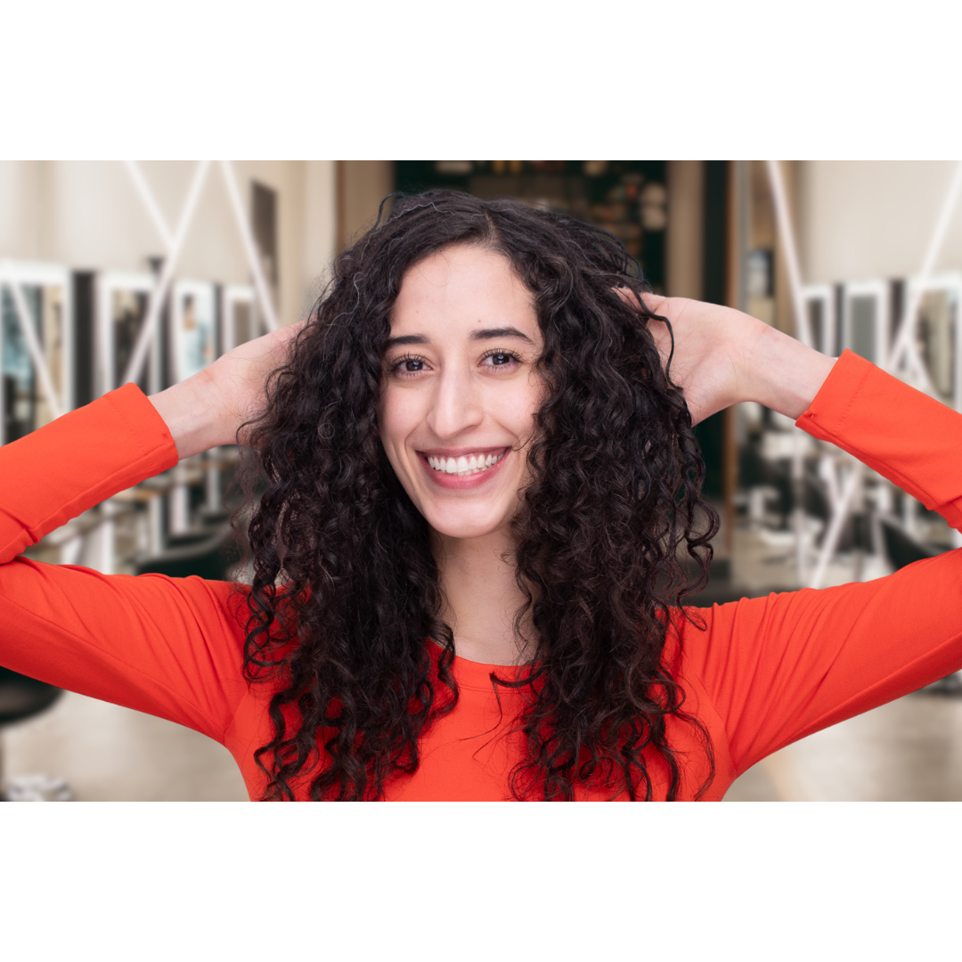 woman in red shirt with brown curly hair smiling, with her hand behind her back, showing off the volume of her hair