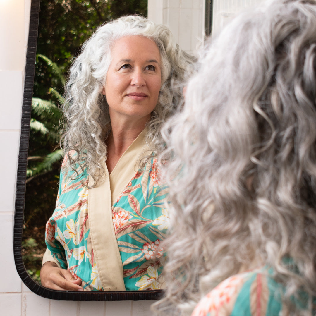 gray, curly haired woman smiling and looking into the bathroom mirror with foliage around