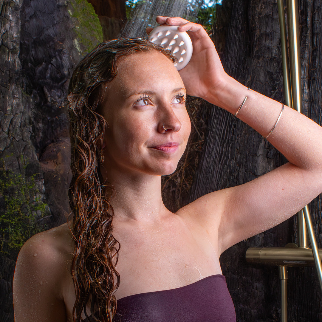  curly haired woman in a stump shower surrounded by redwood trees as she washes her hair using a scalp brush