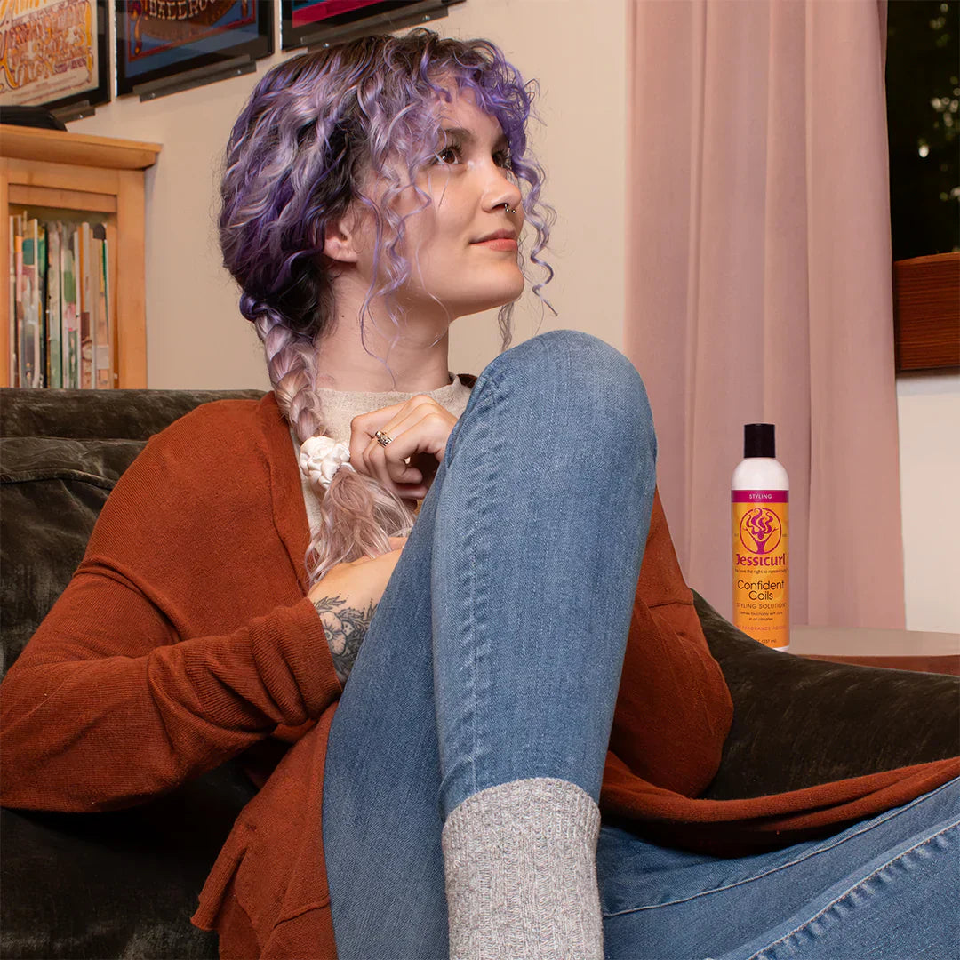 woman with her curly hair in a braid, sitting on the couch with a bottle of Confident Coils Styling Solution next to her