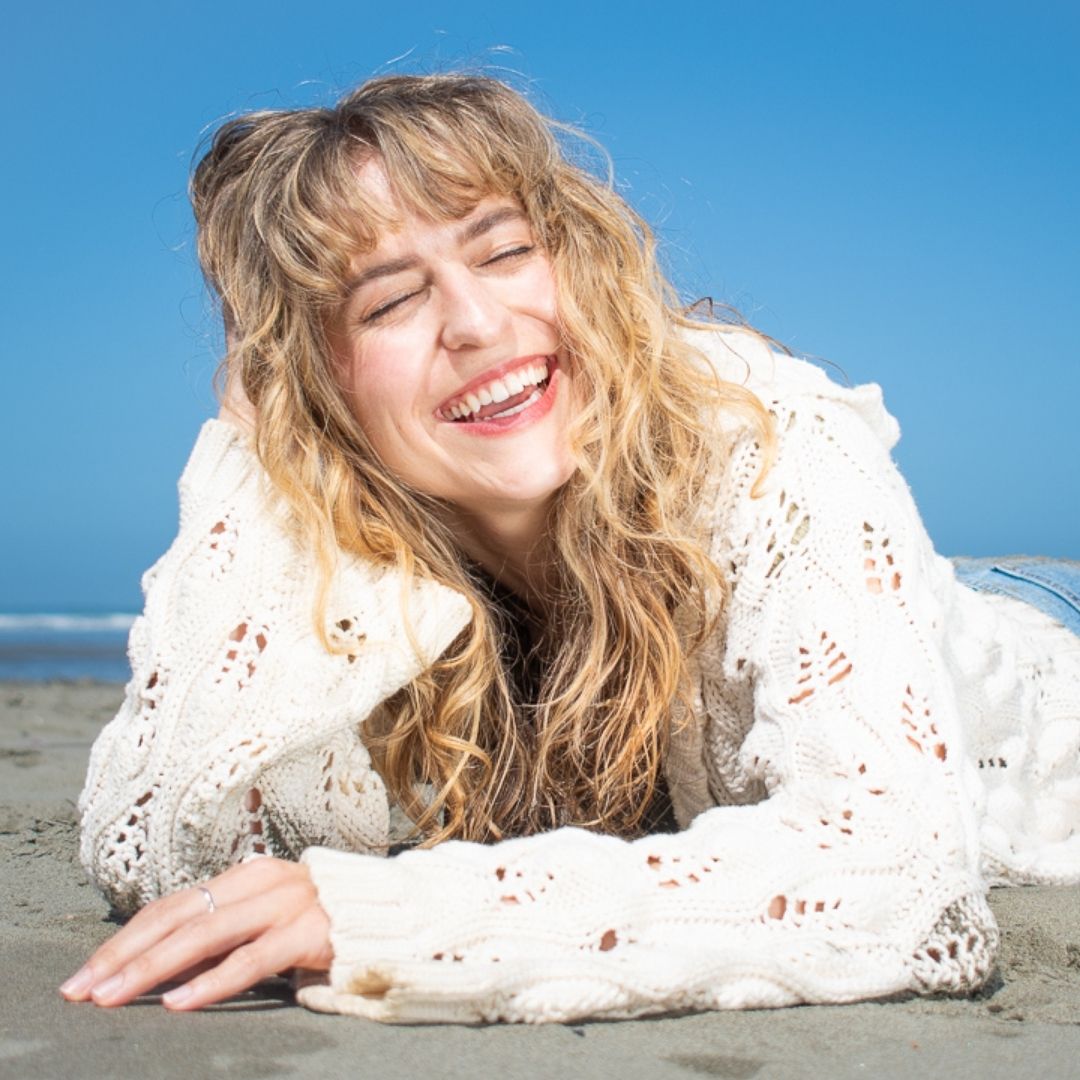 smiling , blonde, curly haired woman laying on a sandy beach with her head propped up in her hand