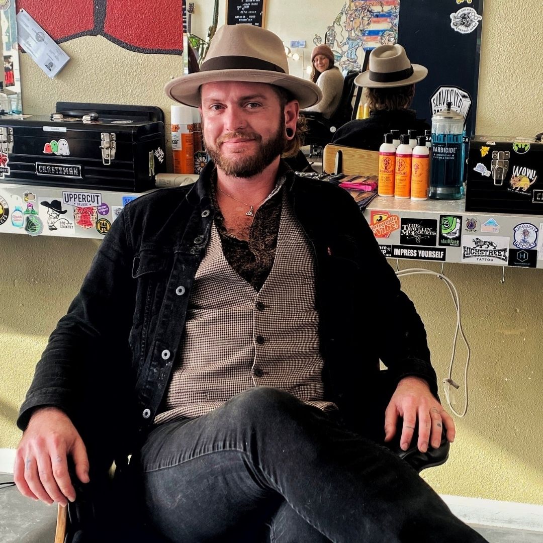 curly haired barber in hat and casual suit sitting at his own chair at his work station