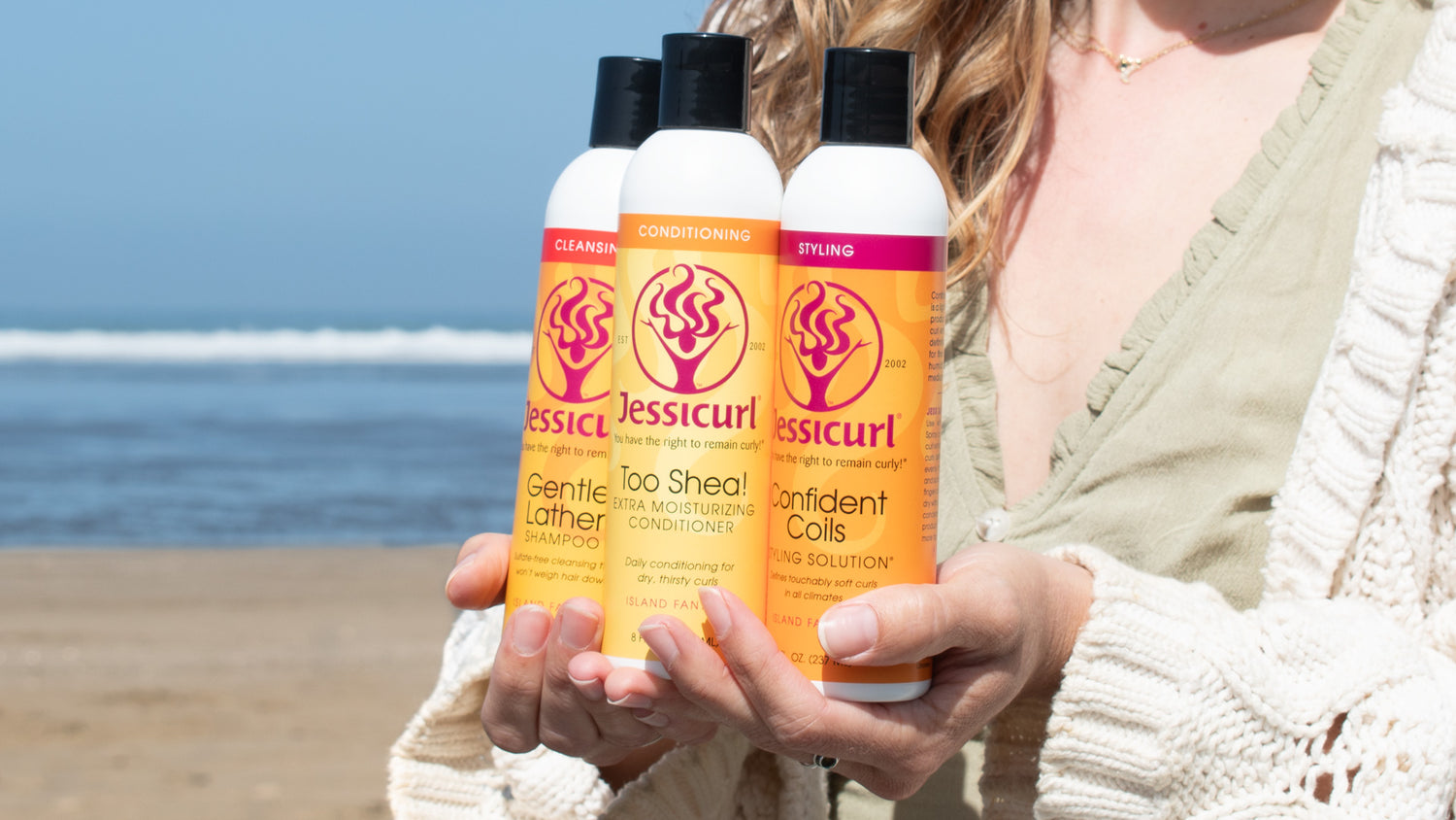 Woman with wavy hair holding a Jessicurl cleanser, conditioner, and styler for her hair type on beach.