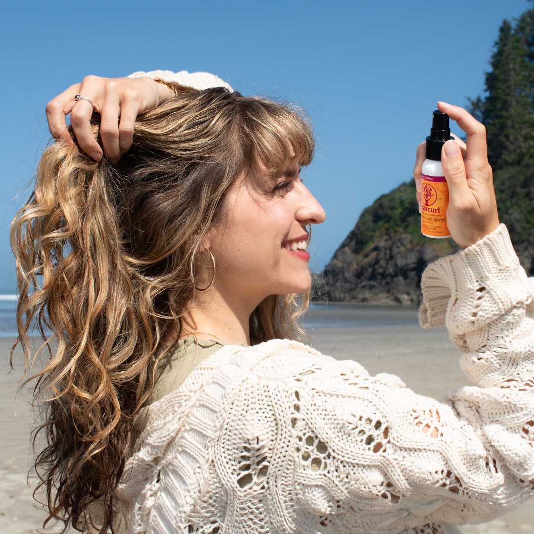 smiling curly haired woman on the beach sectioning her hair with one hand and applying Gelebration Spray 