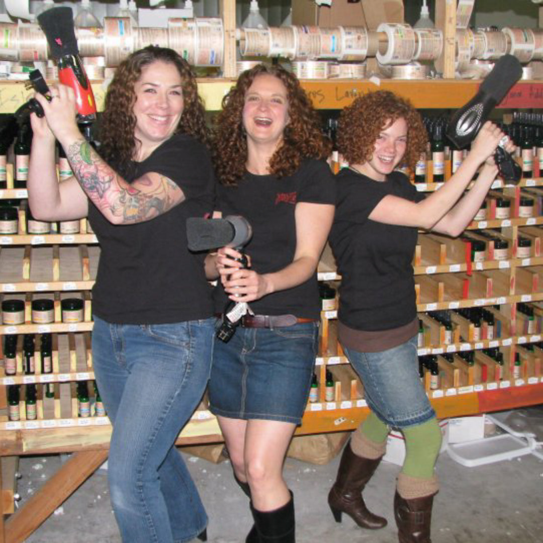 2002 - Photo of Jess (middle), standing with employees in front of label & bottle racks in warehouse, recreating the Charlie's Angel pose using hair dryers with diffusing socks on the end.